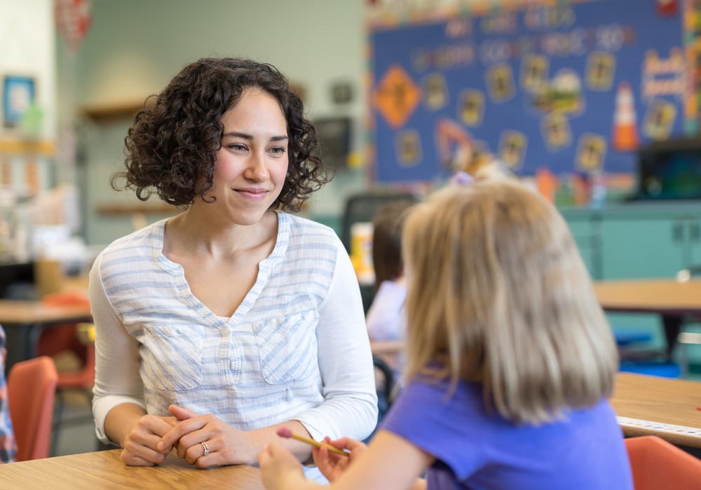 A young girl looks up from her desk to talk with her teacher in a classroom setting